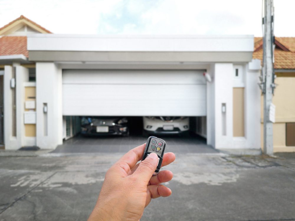 The Pros And Cons Of Automatic Vs. Manual Garage Doors 1 first person view of a hand with a garage door remote and in the back a garage door opening